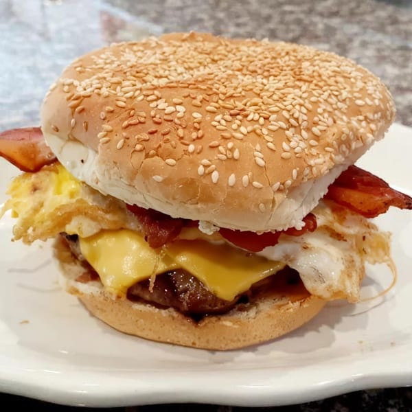 A breakfast burger with a sesame seed bun, bacon, fried egg, and melted cheese on a white plate.